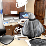 Dental treatment room with patient chair, assistant stool, computer monitor, and overhead dental equipment
